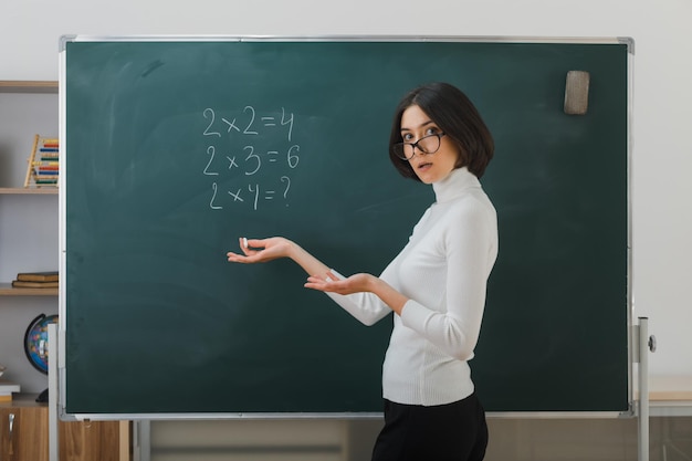 confused spreading hands young female teacher wearing glasses standing in front blackboard and writes in classroom