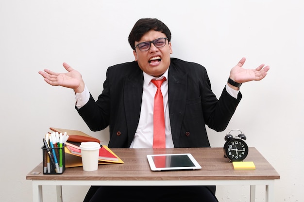 Photo confused stressed asian businessman sit work at office desk spread hands shrugging shoulders isolated on white background