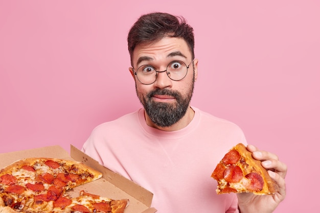 Confused surprised unshaven young man eats tasty pizza has fast food addiction enjoys tasty snack in pizzeria looks wondered, poses against pink wall. Unhealthy nutrition concept