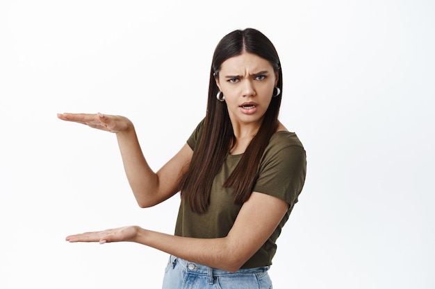 Confused woman holding empty copy space for logo or banner, showing big size box gesture and stare puzzled at front, standing against white wall