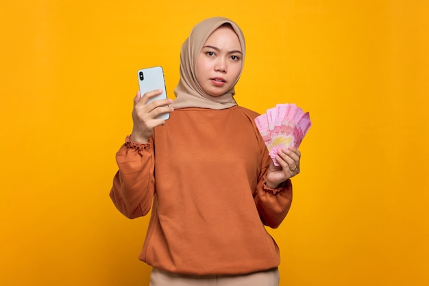 Confused young Asian woman in orange shirt holding mobile phone and showing money banknotes isolated over yellow background