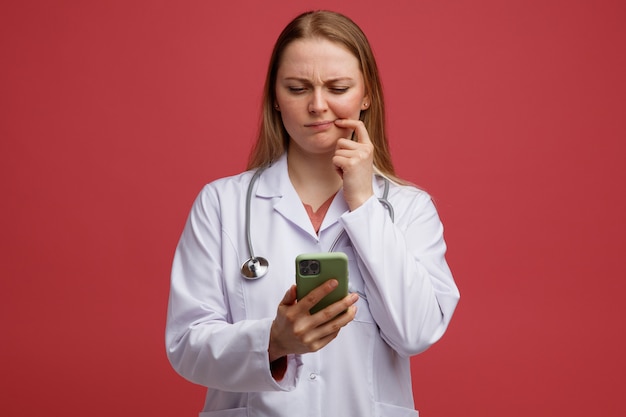 Confused young blonde female doctor wearing medical robe and stethoscope around neck holding and looking at mobile phone keeping finger on corner of mouth 