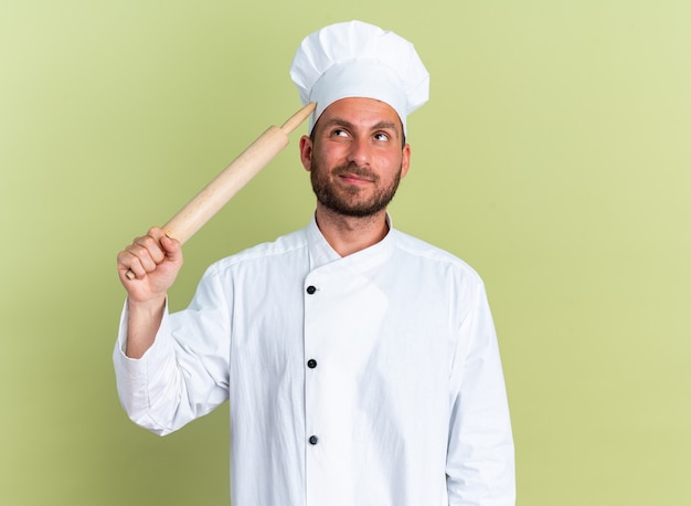 Confused young caucasian male cook in chef uniform and cap touching head with rolling pin looking up isolated on olive green wall