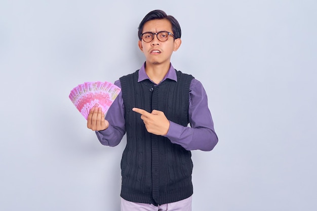 Confused young handsome Asian man in shirt clothes pointing finger at cash money in rupiah banknotes in hands isolated on white background People lifestyle concept