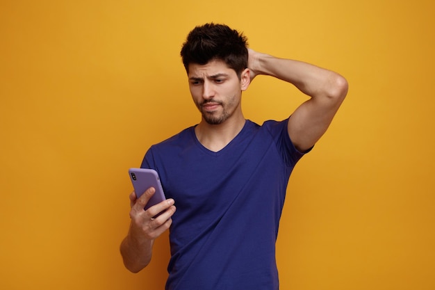 Confused young handsome man holding and looking at his mobile phone keeping another hand on back of his head on yellow background