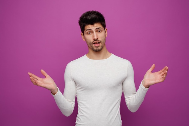 Confused young handsome man looking at camera showing empty hands on purple background