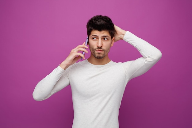 Confused young man keeping hand on back of head looking at side while talking on phone isolated on purple background