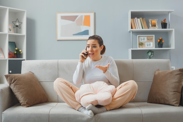 Confused young woman in casual clothing sitting on a couch at home interior looking displeased while talking on mobile phone raising arm in displeasure