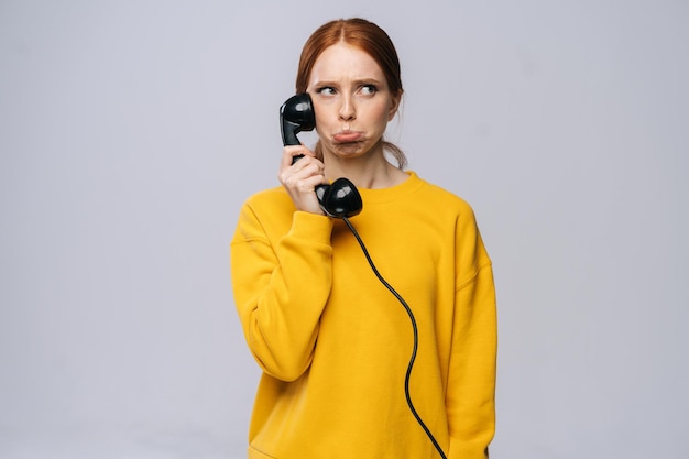 Confused young woman in stylish yellow sweater talking on retro phone and looking away