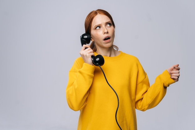 Confused young woman in stylish yellow sweater talking on retro phone and looking away