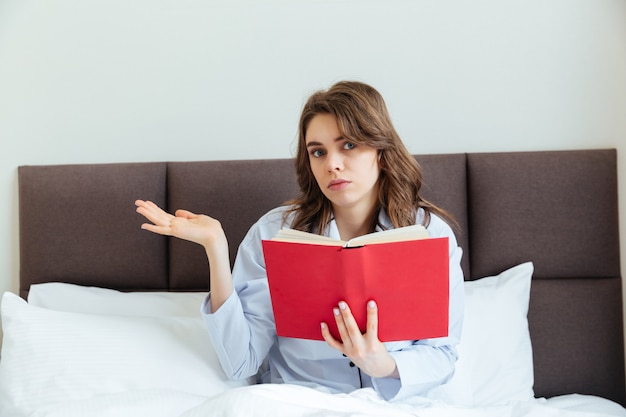 Confused young woman wearing pajamas and holding book in bed