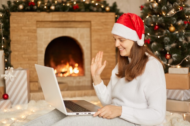 Congratulations via Internet. Smiling girl at home using laptop computer notebook for video call, woman looking camera and waving greeting hands, being happy, sitting on floor with laptop on knees.