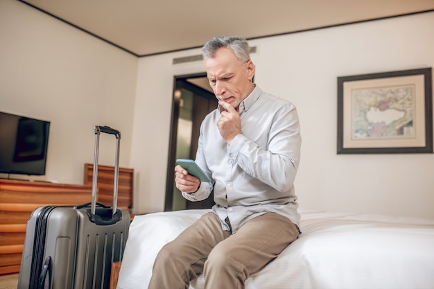 Connected. Mid-aged businessman sitting on a bed in a hotel with a smartphone in hands