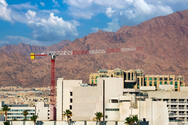 Construction crane against mountains range with fluffy clouds Modern house under construction in a new area of high buildings on a background of mountains in Eilat Israel Red construction crane