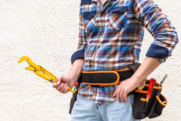 Photo a construction worker in a blue checked shirt with tools in his belt holds a yellow wrench in his hands.