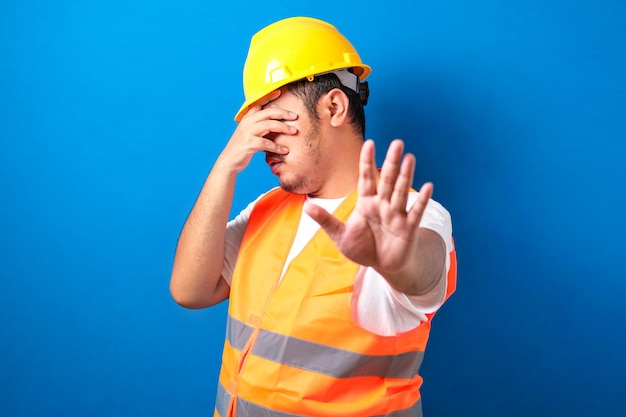 construction worker man wearing uniform and helmet covering eyes with hands and doing stop gesture