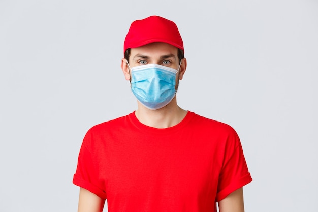 Contactless delivering, covid-19 and shopping concept. Close-up of young man courier in red uniform, cap and medical mask looking at camera, advertisement of deliver company