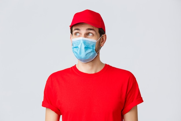 Contactless delivering, covid-19 and shopping concept. Thoughtful courier in red uniform cap, t-shirt and medical face mask with gloves, looking upper left corner, reading banner or advertisement