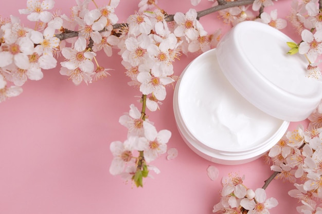 Container with bodycare and skincare cream on a pink background with blooming cherry Cosmetic facial skin care and spa Natural treatment concept