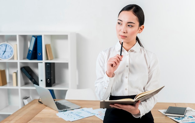 Contemplated young asian businesswoman standing in front of desk holding diary and pen in hand