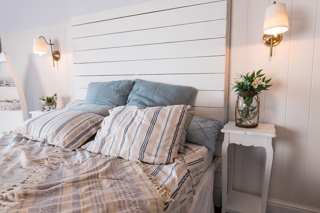 Contemporary Bed dressed with pastel colored cushions and handing lamp.Vase with spring flowers standing on white sidetable.bright bedroom interior. Comfortable bed with coverlet and striped pillows.