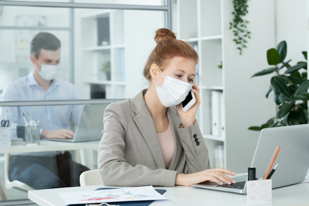 Contemporary businesswoman in protective mask looking at laptop display while working with data and phoning in office against her colleague