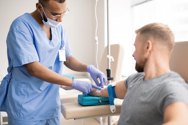 Contemporary doctor in protective gloves and mask preparing arm of sick patient for dropper injection in hospital