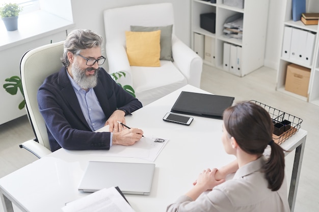 Contemporary employer in formalwear making notes in resumer of new applicant while sitting in armchair by desk in front of her