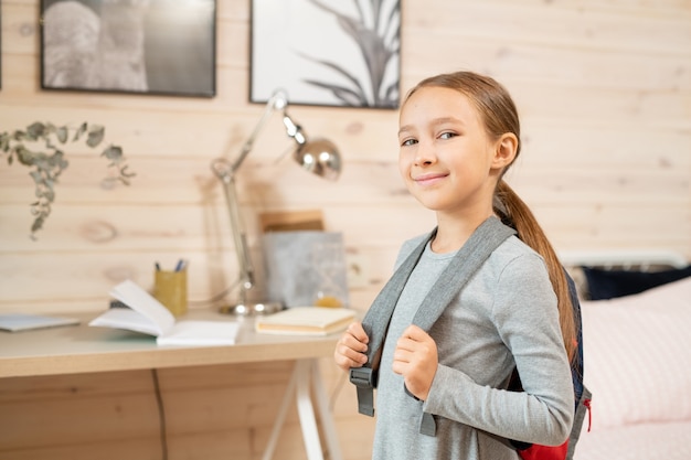 Contemporary happy schoolgirl with backpack standing in front of camera against her bed and table with books by wooden wall with two pictures