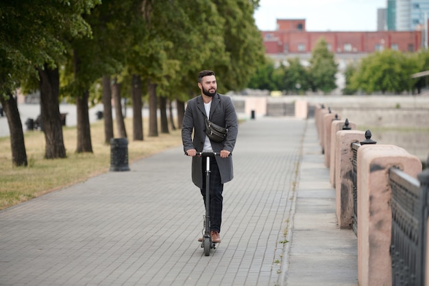 Contemporary young bearded businessman in coat riding on electric scooter in urban environment along riverside in the morning