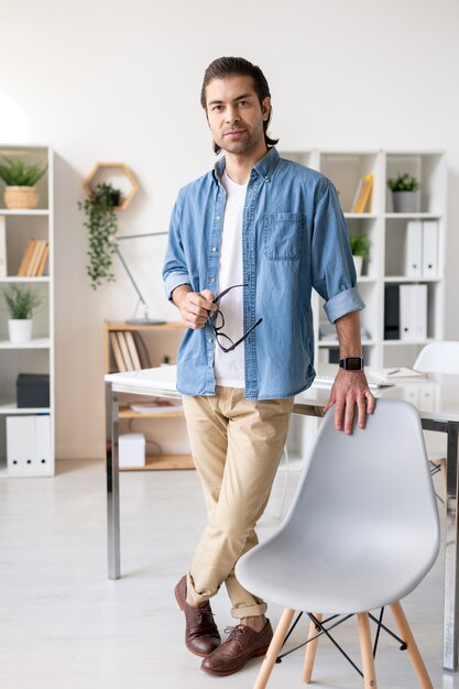 Contemporary young businessman in casualwear standing by chair in front of camera with desk on background