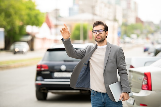 Contemporary young man with laptop standing by road with raised arm while catching taxi in the morning