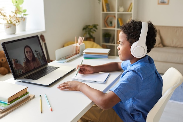 Content AfricanAmerican schoolboy sitting at table and attentively listening to online tutor in headphones