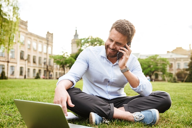 content european man in business clothing, sitting on grass in park with legs crossed and talking on cell phone while using silver laptop