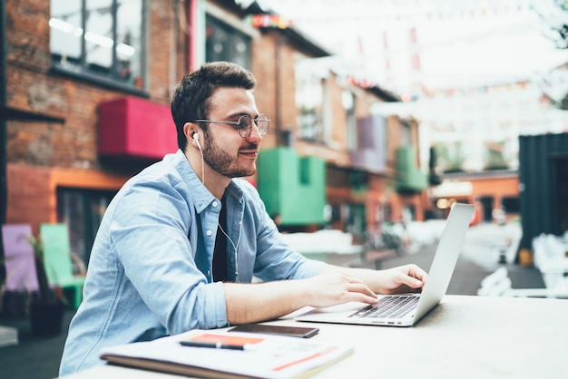 Content man in casual outfit and earphones smiling while browsing video on laptop at cafe