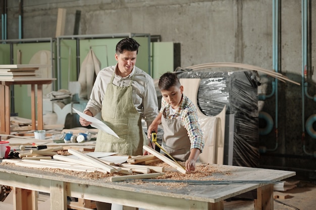 Content satisfied father with sketch looking at teenage son measuring wooden plank in woodworking studio