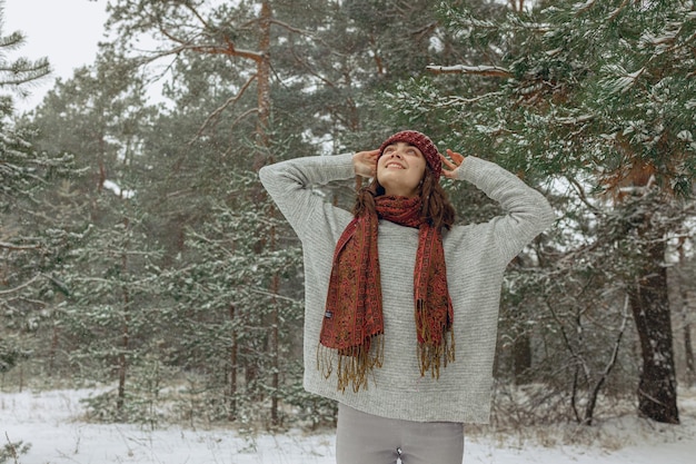 Content woman in knitted sweater warm hat and scarf standing in snowy evergreen forest in winter