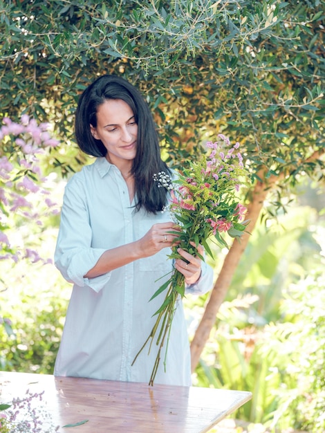 Content woman in white shirt standing at table in park and adjusting flowers in hands in daylight