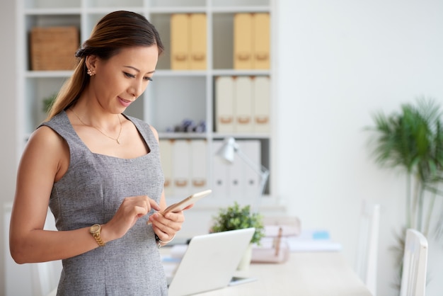 Content young Asian manager in wristwatch reading message on smartphone while standing in office