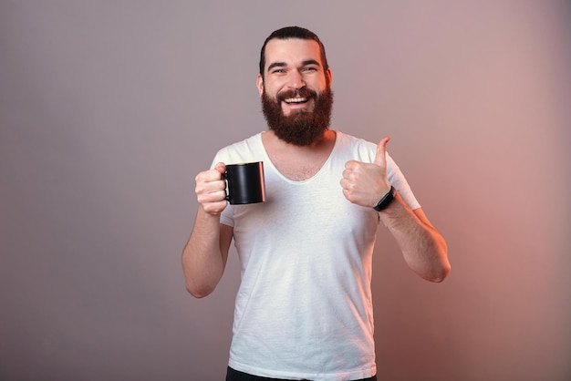 Content young man is holding a black mug while showing thumb up for like