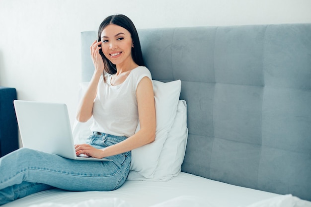 Contented attractive young woman smiling and touching her temple while sitting in a bed with her laptop Template banner