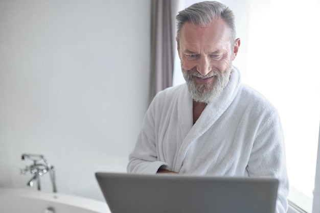 Contented man with the laptop standing in the bathroom