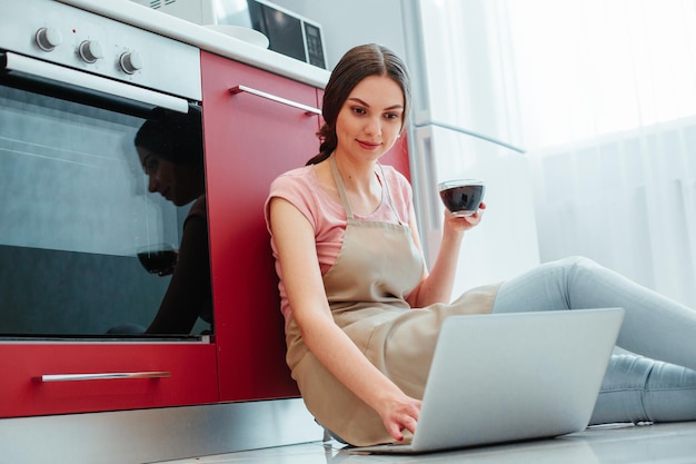 Contented young woman wearing an apron with casual clothes sitting on the floor with coffee and laptop