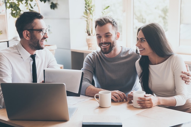 Contract with best conditions! Confident young man in shirt and tie holding some document and pointing it with smile while sitting together with young couple at the desk in office