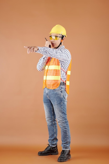 Contractor in bright orange vest and hard hat controlling work at construction site and talking on w...