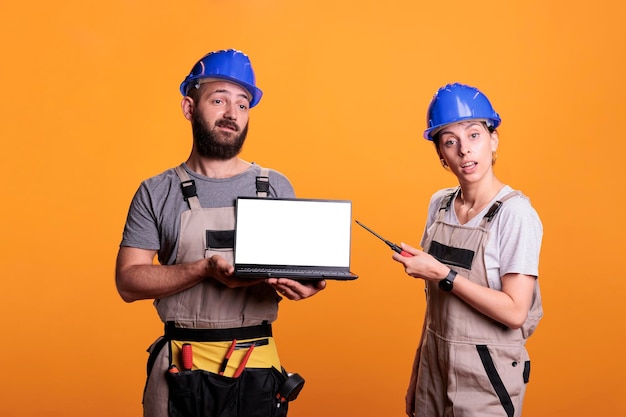 Contractors team showing white blank screen on laptop, wearing overalls and hardhats in studio. Construction workers advertising isolated copyspace display on portable computer, empty template.