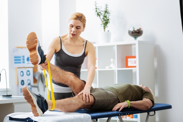 Controlling everything. Beautiful blonde woman looking at her patients leg while controlling the performance of the exercise