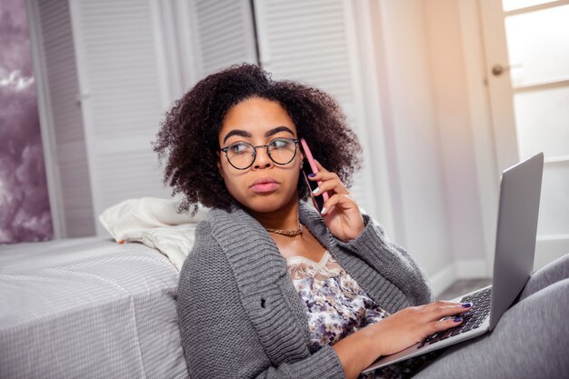 Conversation on phone. Serious young woman wearing clear glasses during working with laptop and talking with smartphone