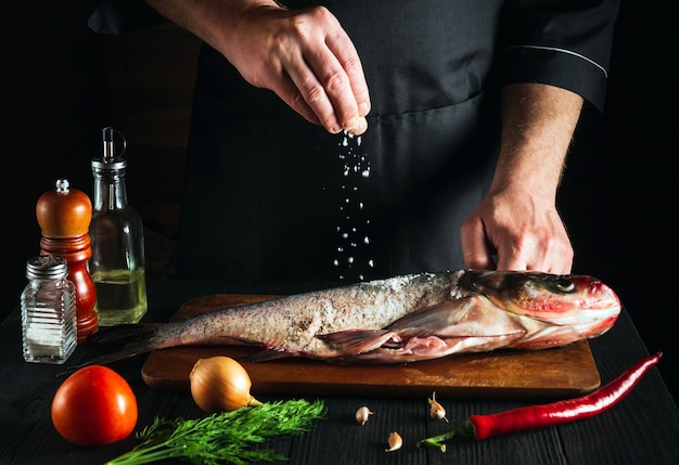 Cook or chef prepares fresh fish bighead carp sprinkling salt with the ingredients. Preparing to cook fish food. Working environment in the restaurant kitchen