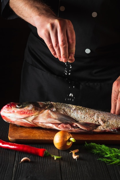 The cook prepares fresh fish bighead carp sprinkling salt with ingredients. Preparing to cook fish food. Working environment in the restaurant kitchen